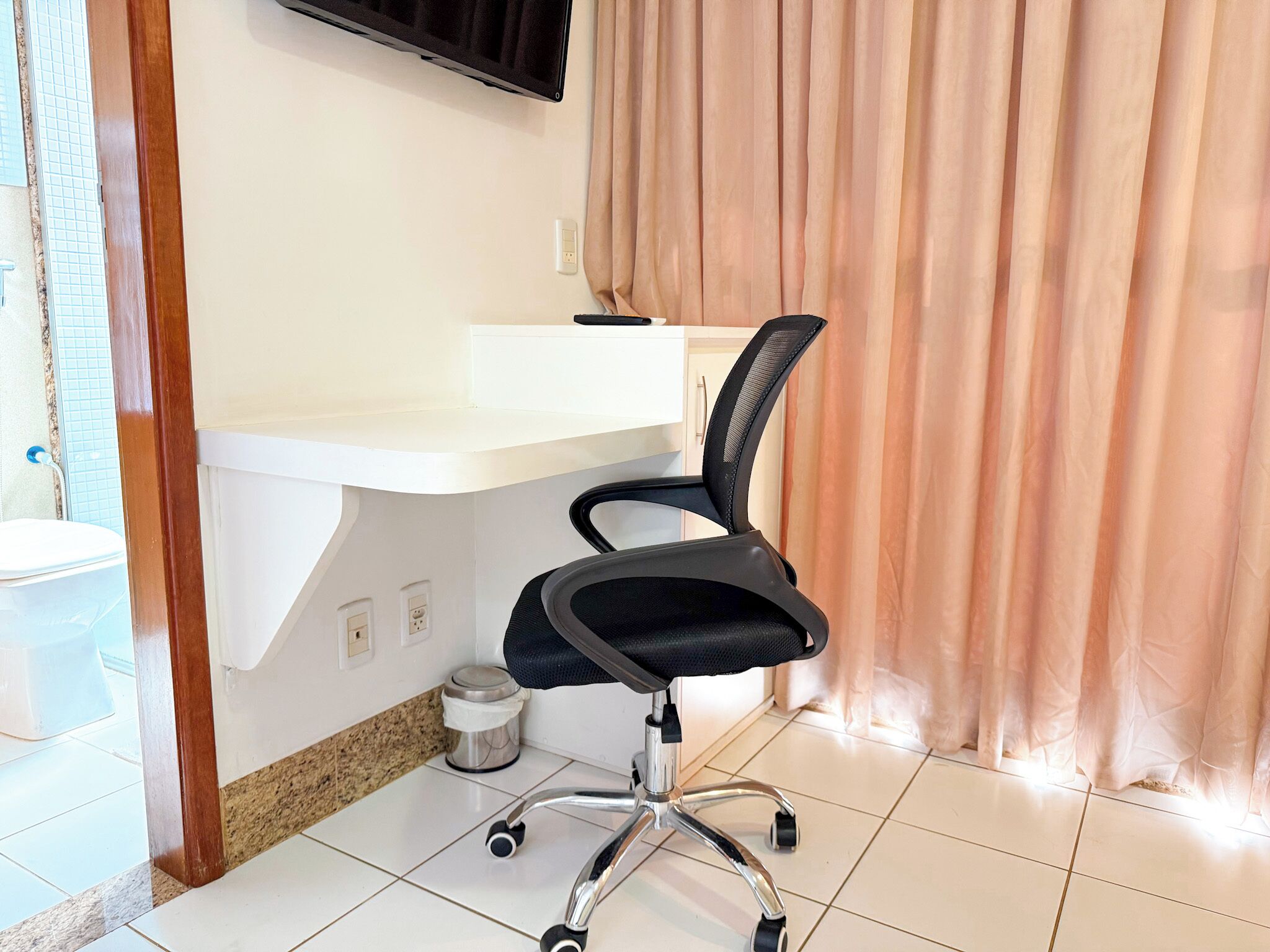 A modern office space with a black office chair, a white desk, and a television mounted on the wall.