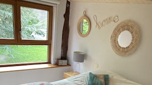 Ein Schlafzimmer mit einem Bett, einer blumigen Tagesdecke, zwei Kissen, einer Nachttischlampe, einem Wandspiegel und einem Fenster mit Blick auf Grünflächen.