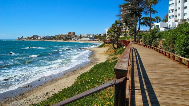 A wooden boardwalk along a beach with ocean views, surrounded by greenery and buildings in the background. - Rocas del Mar I - Front Line Beach (Mijas Costa)