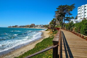 Un paseo marítimo de madera bordeando una playa, con vistas al océano, rodeado de vegetación y edificios al fondo.