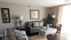 A living room with a sofa, armchair, ottoman, and coffee table. There is a framed picture on the wall, a lamp, and a plant. - Yacht Club S #1-1104 4 Bedroom Condo by RedAwning (North Myrtle Beach)