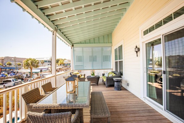 A covered outdoor deck with wicker furniture, a glass table, and a view of the street. - Two Homes w/ Private Pools, Perfect for Wedding Groups (Folly Beach)