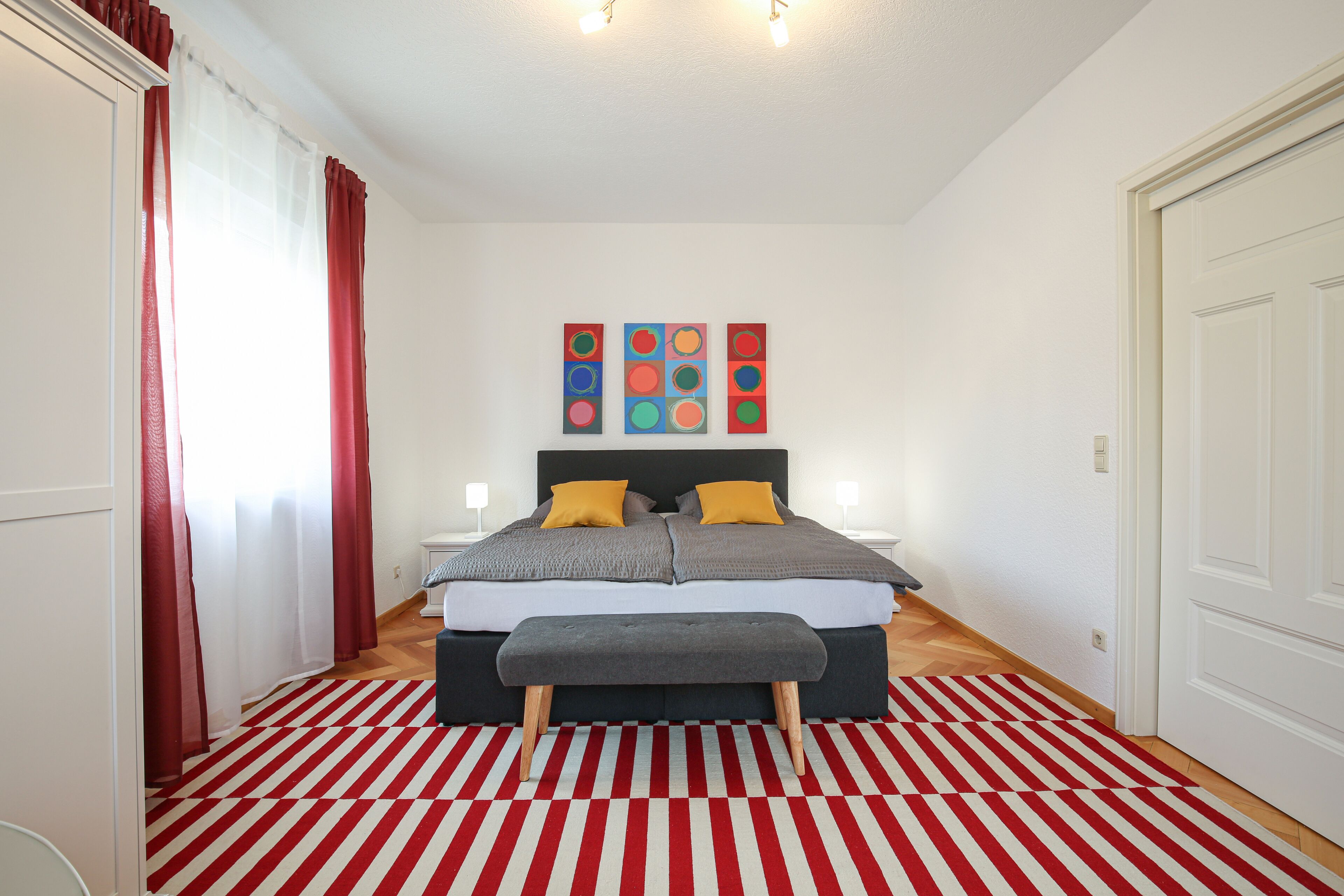 A bedroom with a bed, a bench, a red and white striped rug, and a wall with colorful artwork.