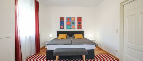 A bedroom with a bed, a bench, a red and white striped rug, and a wall with colorful artwork.