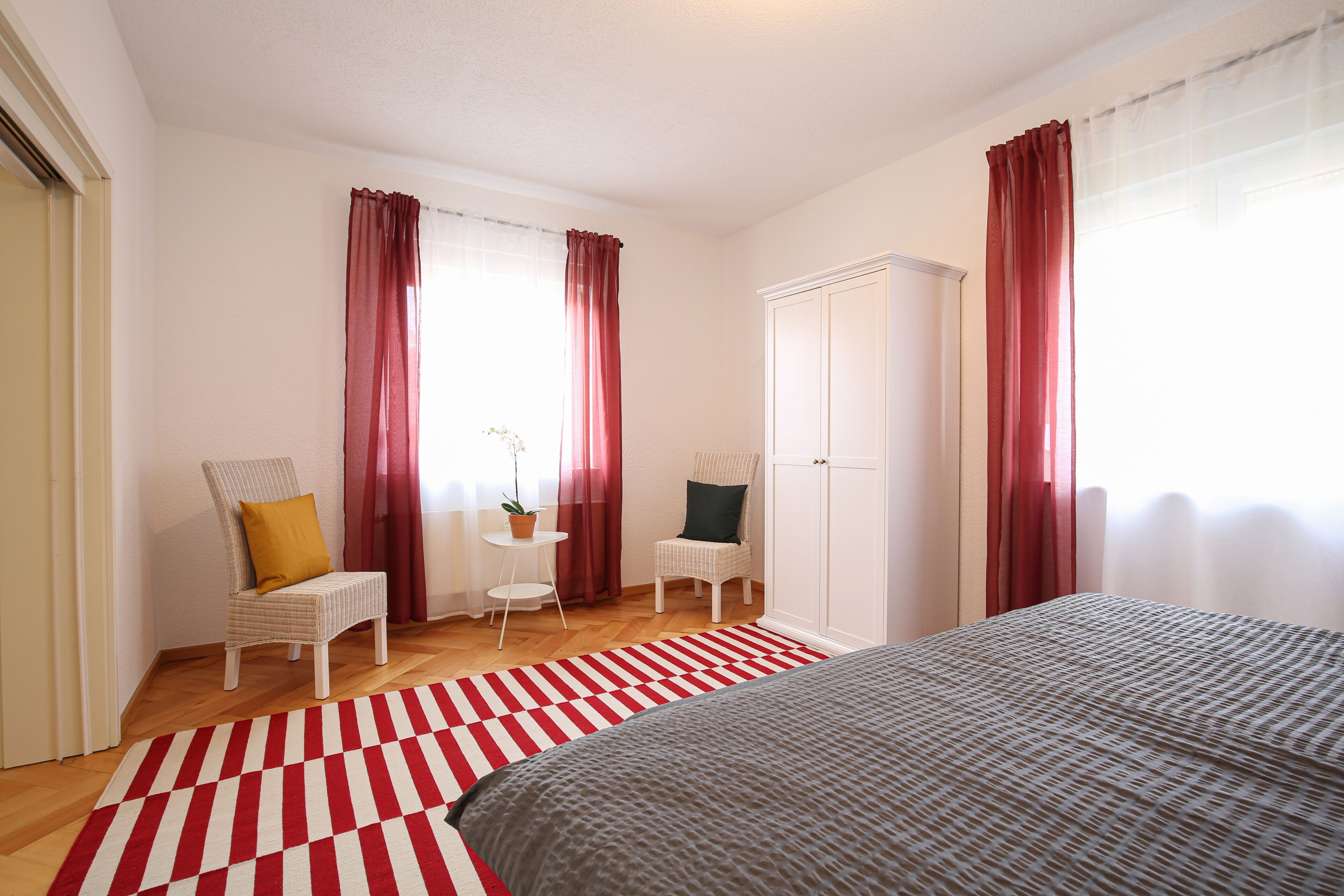 A bedroom with a bed, two chairs, a wardrobe, and a red and white striped rug.
