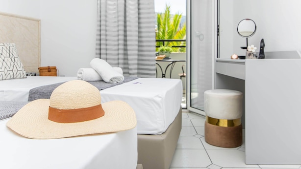 A hotel room with a bed, a large straw hat, a small round mirror, and a view of a balcony with plants. - ICandy Sol (Hersonissos)