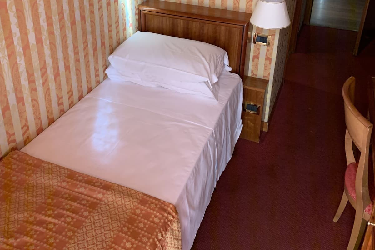 A hotel room with a bed, a nightstand with a lamp, a wooden chair, and a patterned carpet.