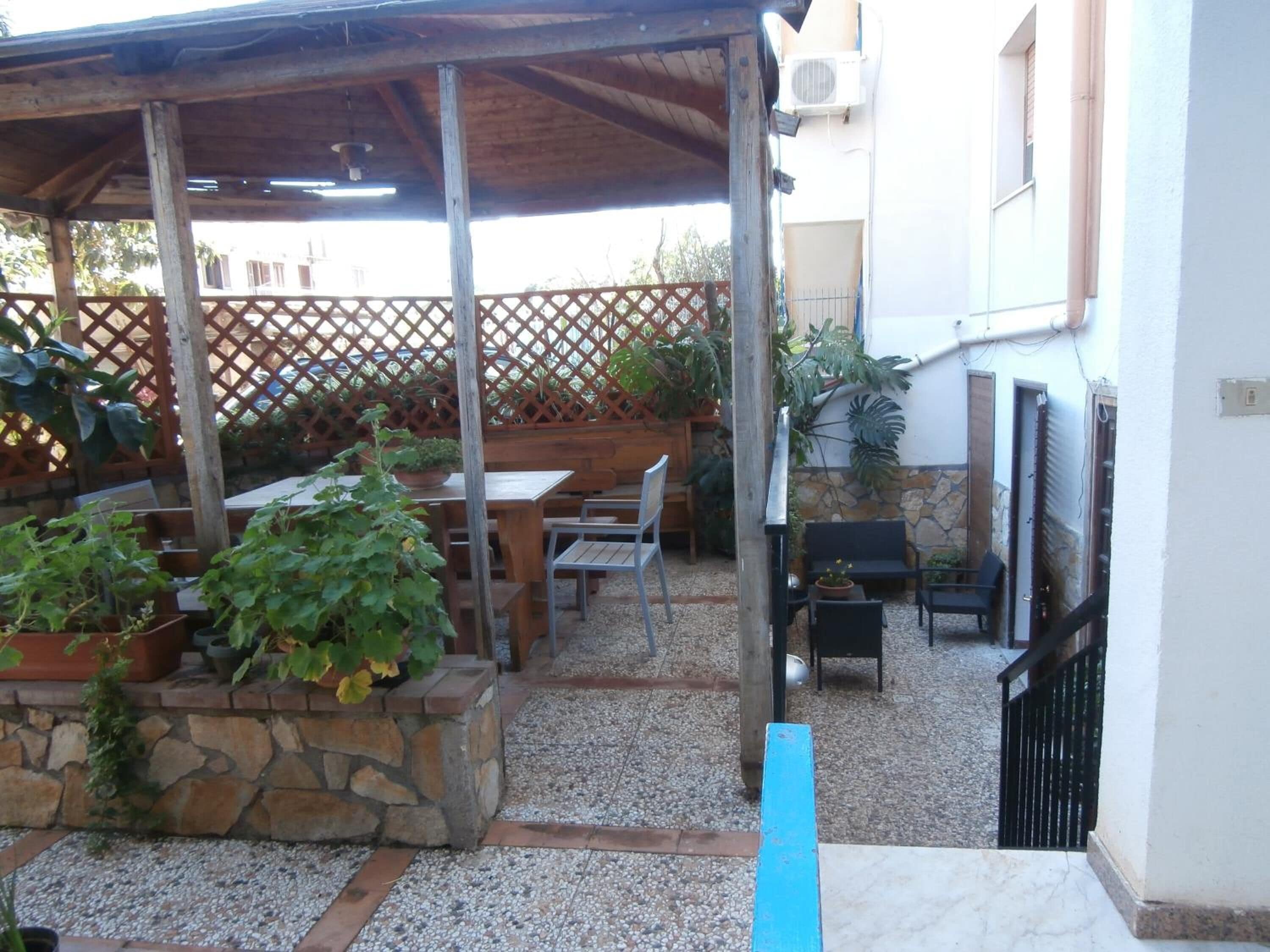 Un'area salotto all'aperto riparata, con tavoli e sedie, circondata da piante in vaso e un recinto in legno.
