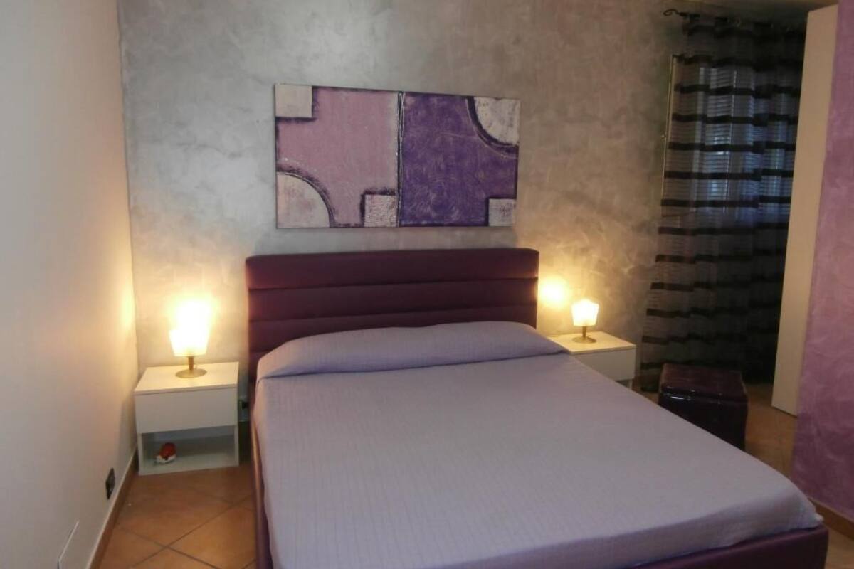 A bedroom with a bed, two bedside tables with lamps, a wall-mounted abstract art piece, and a window with blinds.