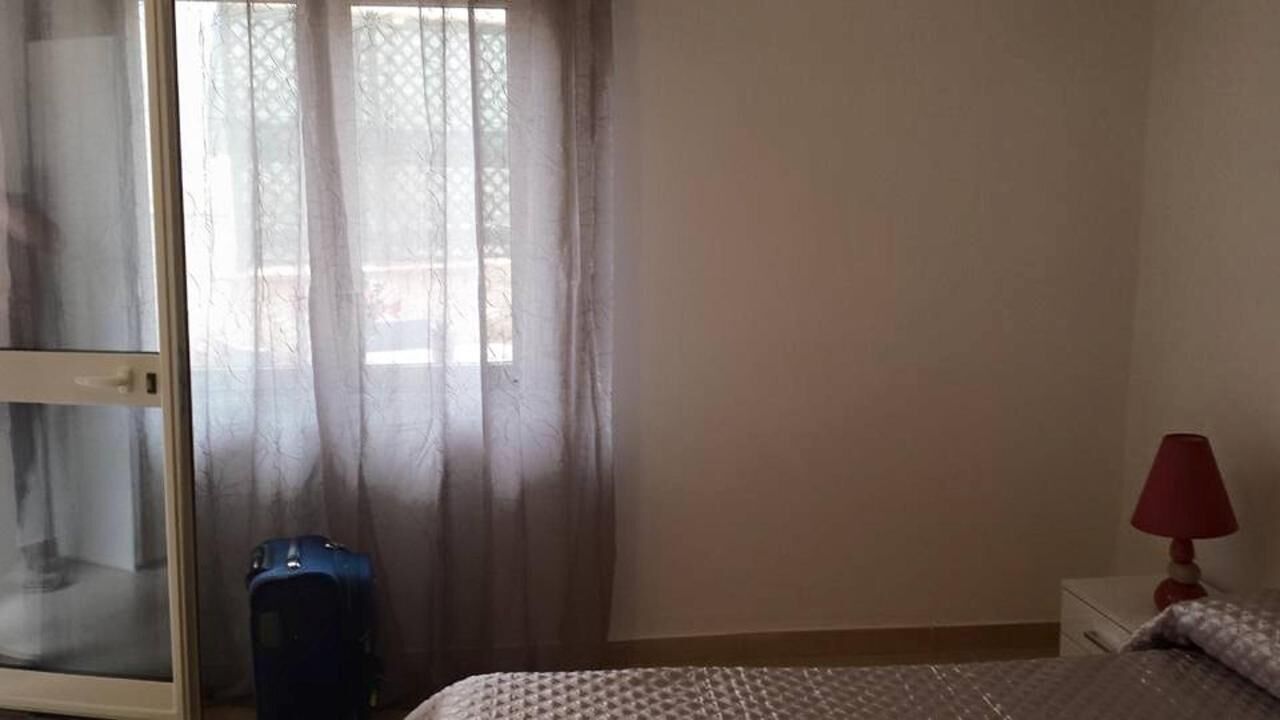 Ein Hotelzimmer mit einem Bett, einem Nachttisch mit Lampe, einem Koffer und einem Fenster mit durchsichtigen Vorhängen.
