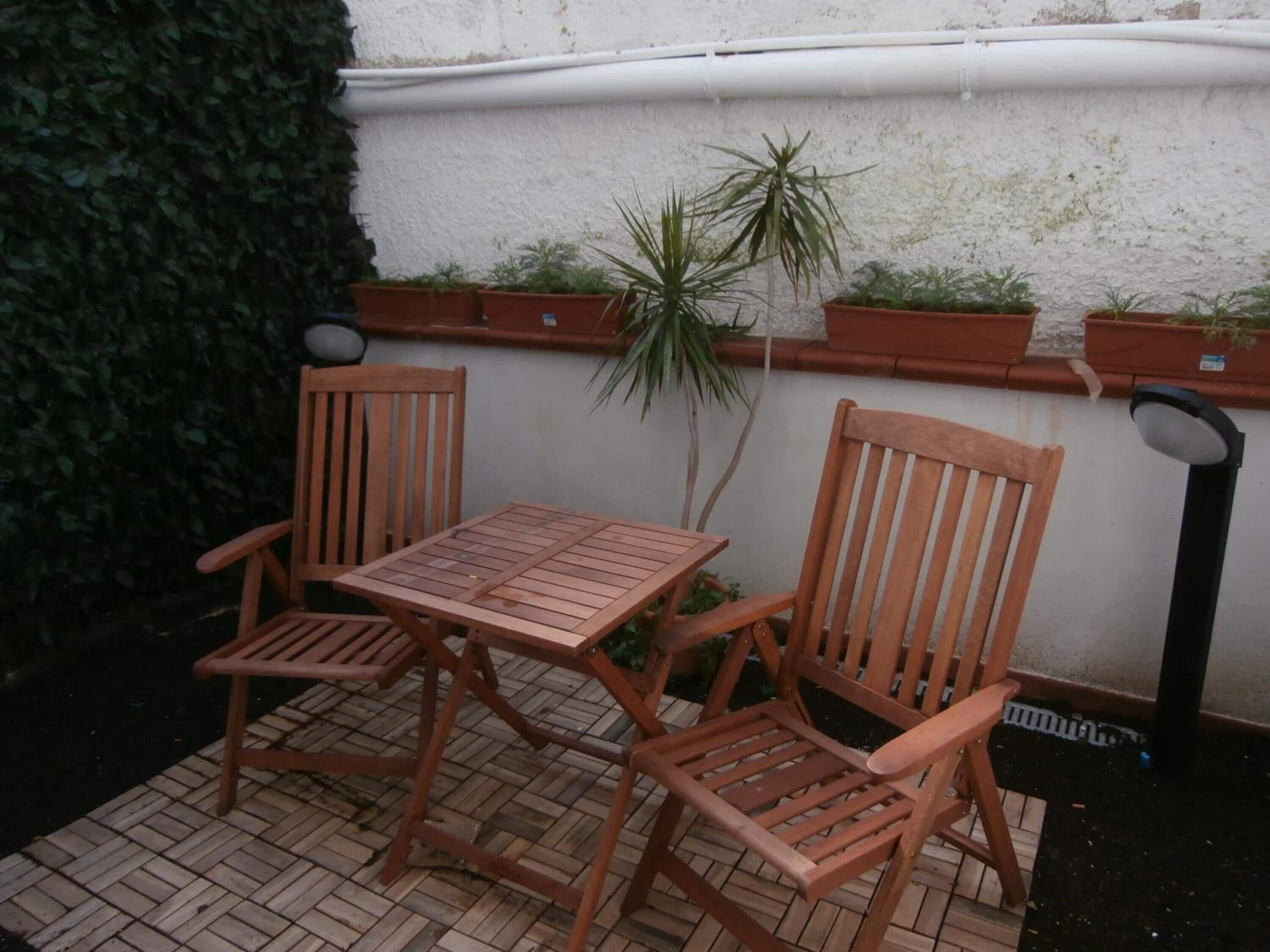 Mesa de madera y dos sillas en un patio al aire libre con piso de baldosas, rodeado de plantas en macetas y una pared blanca.