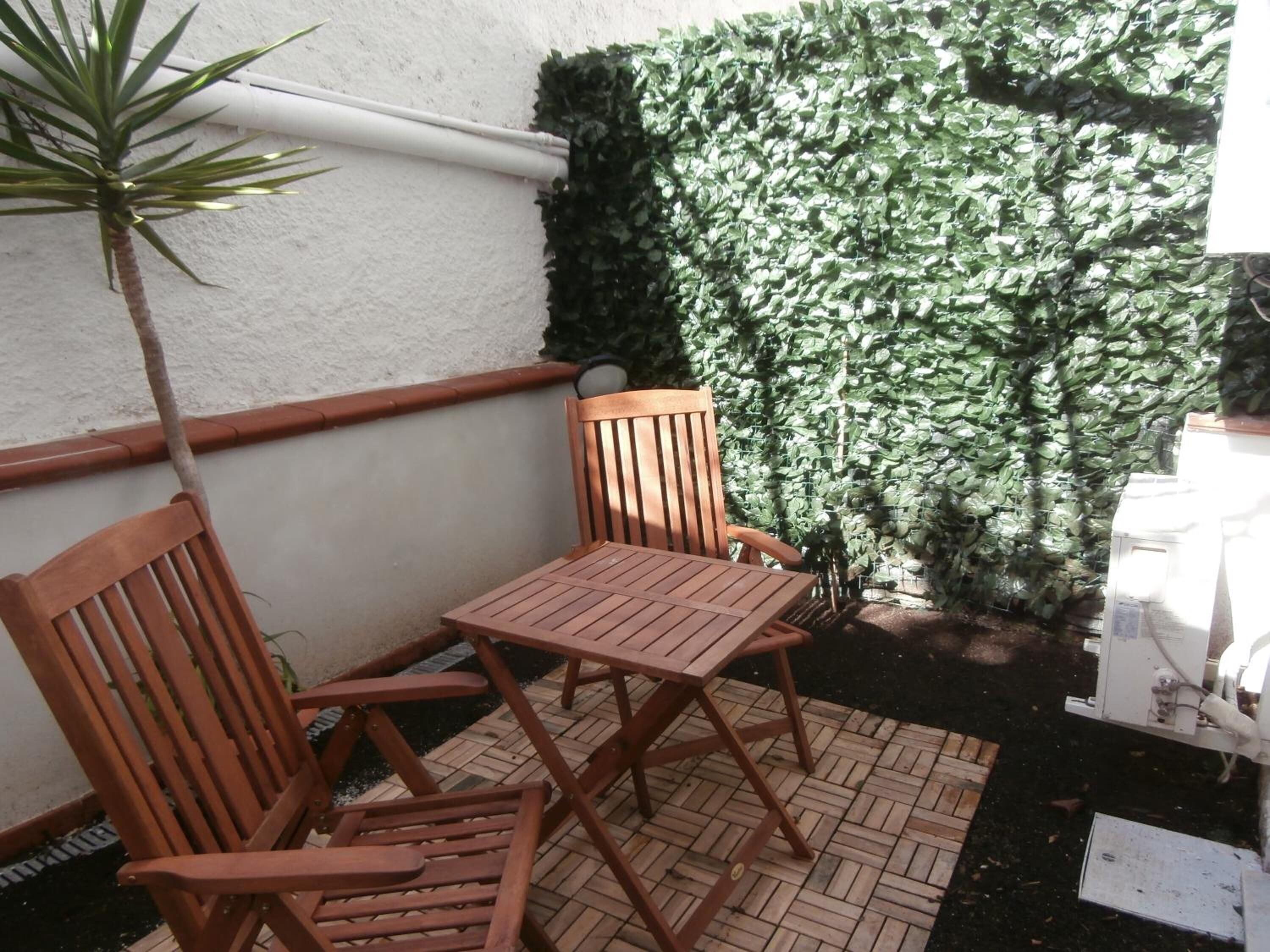 A small outdoor seating area with two wooden chairs and a table, surrounded by a white wall and a green ivy-covered wall.