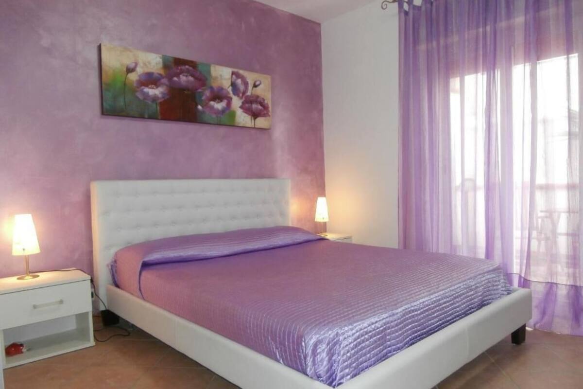 A bedroom with a white bed, purple bedding, a nightstand with a lamp, and a painting on the wall.