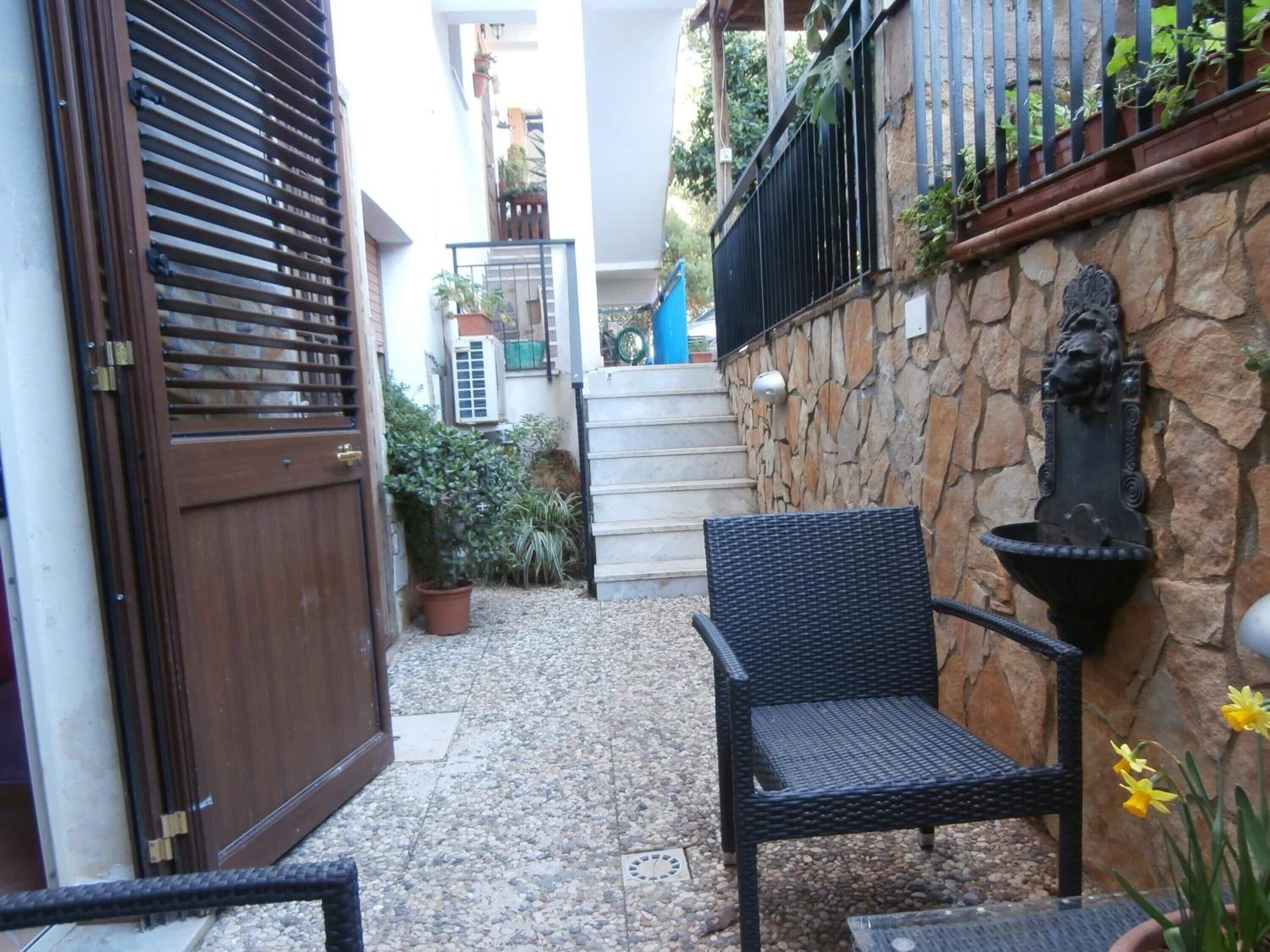 A narrow outdoor area with a stone wall, a black wicker chair, a potted plant, and a decorative wall-mounted fixture.