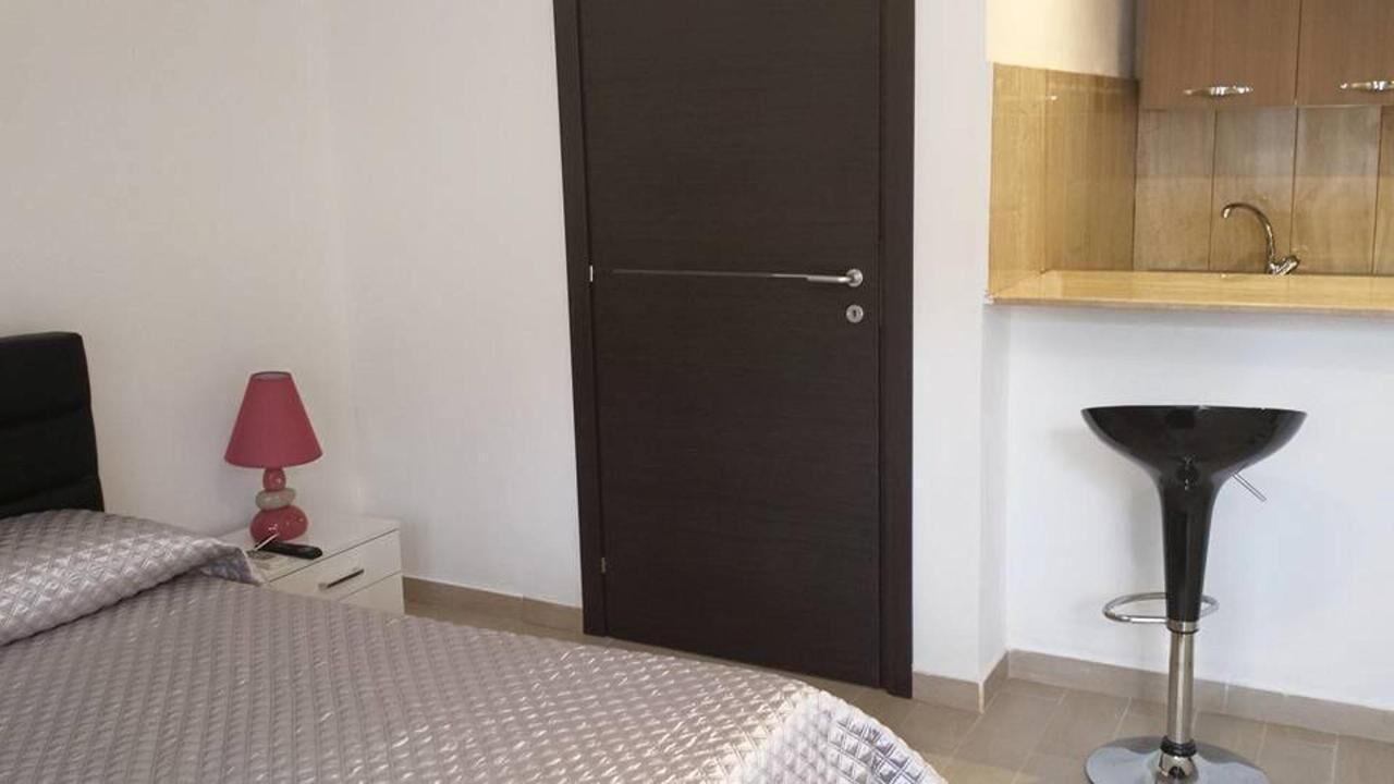 A bedroom with a bed, a nightstand, a lamp, a door, and a kitchen area with a sink.