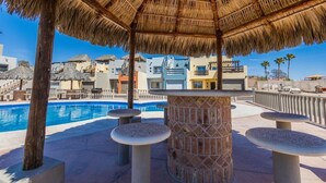 Zona junto a la piscina con una sombrilla de paja, asientos de piedra y vista a edificios al fondo.