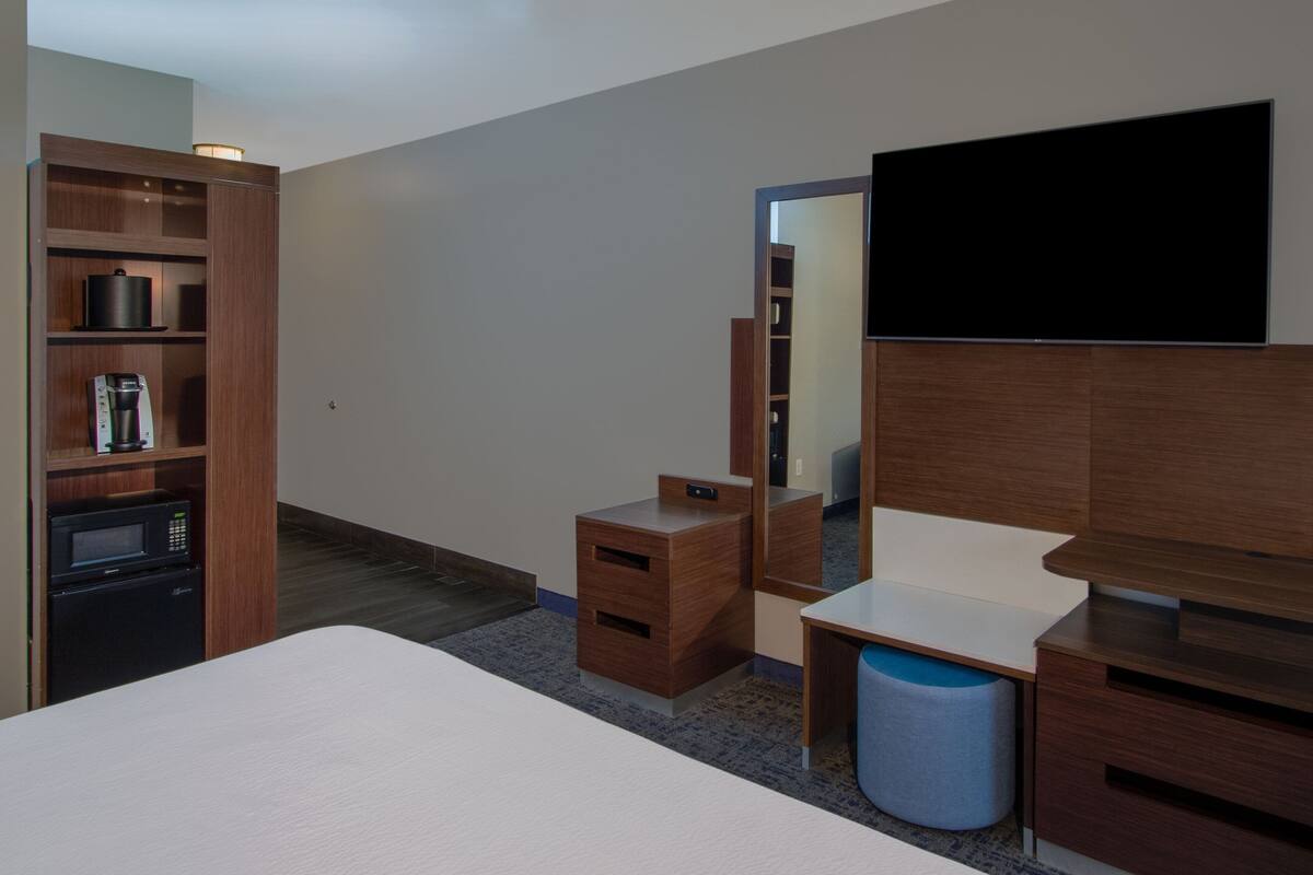 A modern hotel room with a bed, a desk, a wardrobe, a microwave, a coffee maker, and a television.