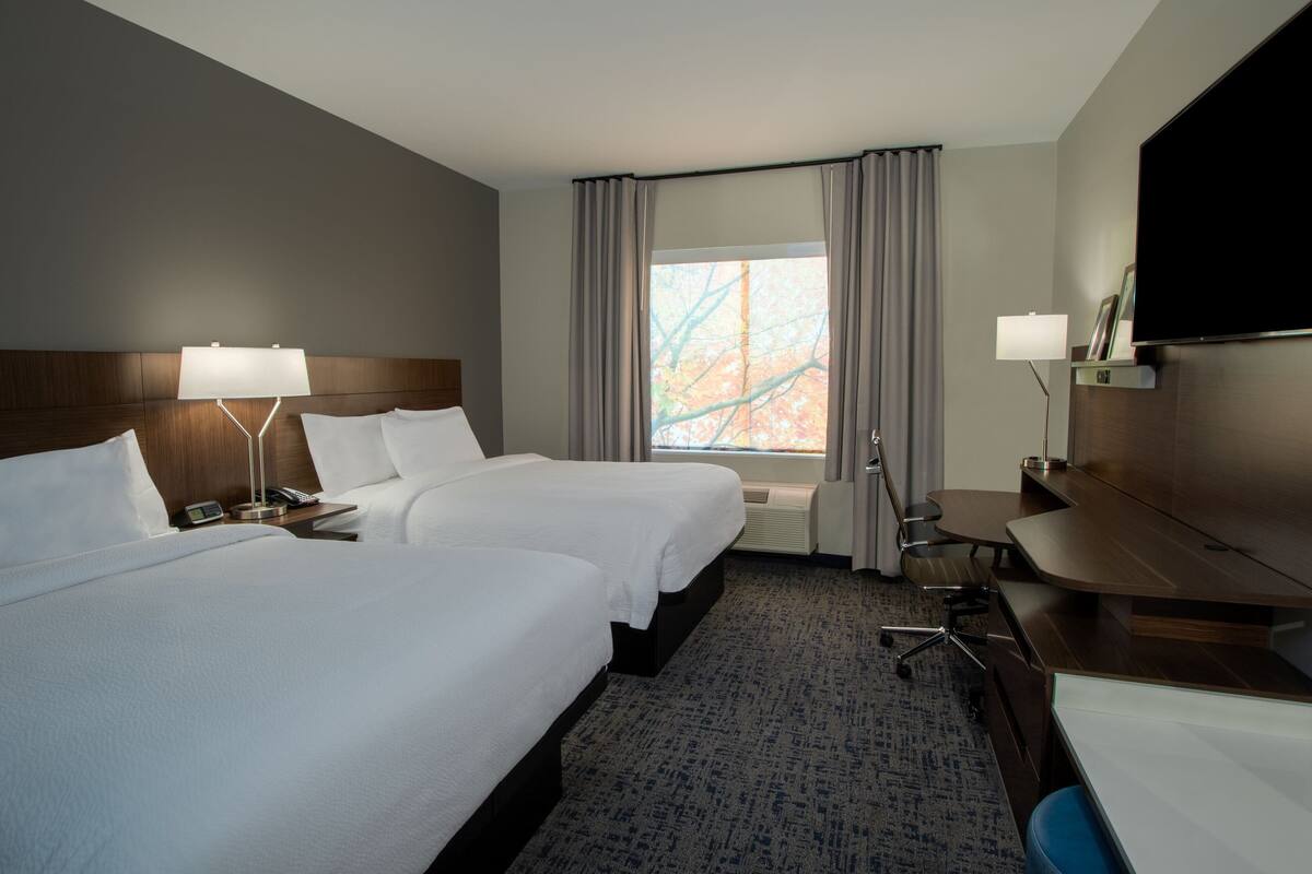 A hotel room with two beds, a desk, a chair, a TV, and a window with curtains.