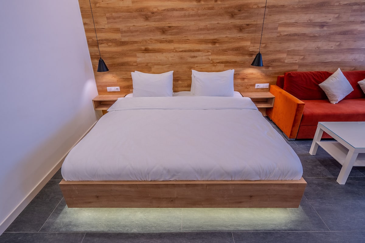 A modern bedroom with a wooden headboard, a bed with white bedding, two bedside tables with lamps, and a red sofa.
