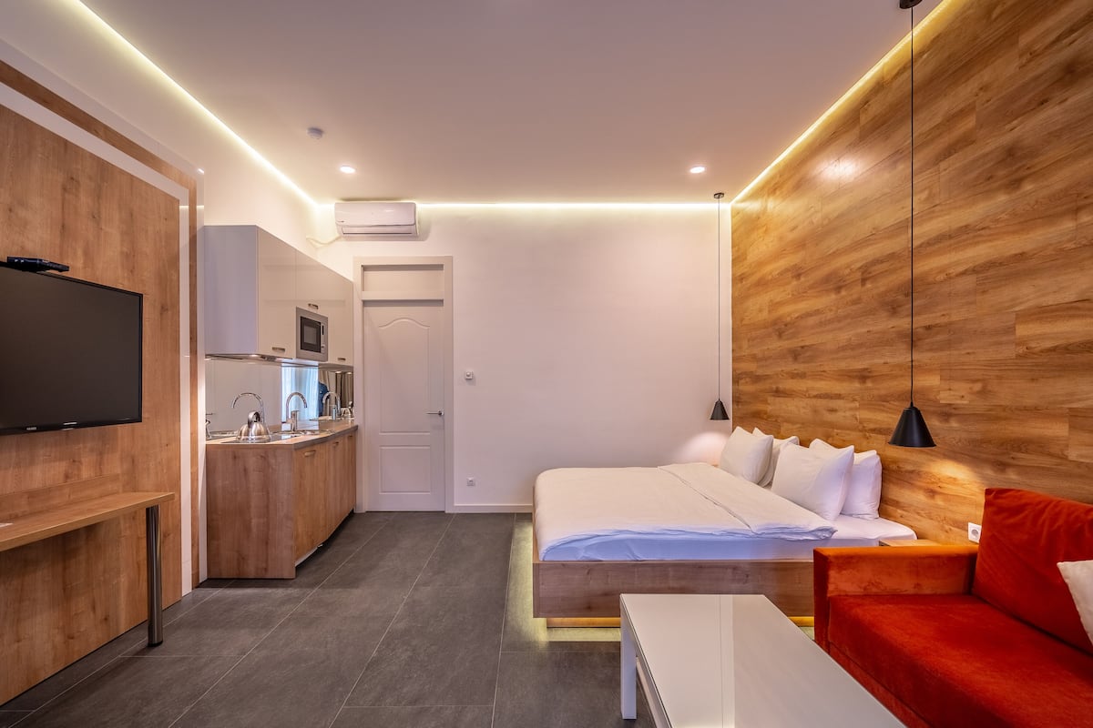 A modern hotel room with a wooden accent wall, a bed with white bedding, a red sofa, a small kitchen area with a microwave, and a flat-screen TV.