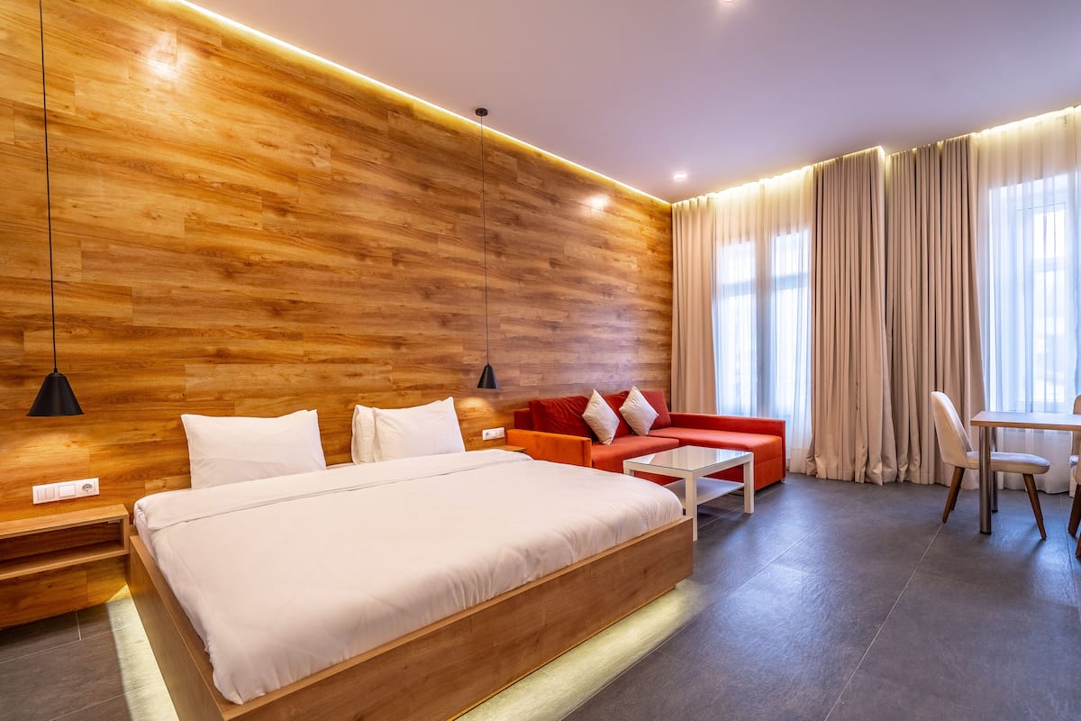 A modern hotel room with a wooden accent wall, a large bed, a red sofa, a glass coffee table, and a dining area with a table and chairs.