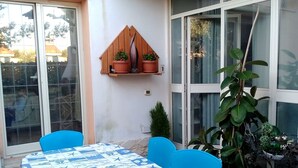A patio area with a table covered by a blue and white patterned tablecloth, two blue chairs, a wooden shelf with potted plants, and a large green plant.