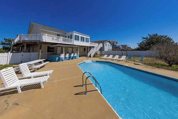 Ein Swimmingpool mit Liegestühlen, einer überdachten Terrasse und einem zweigeschossigen Haus mit Balkon.