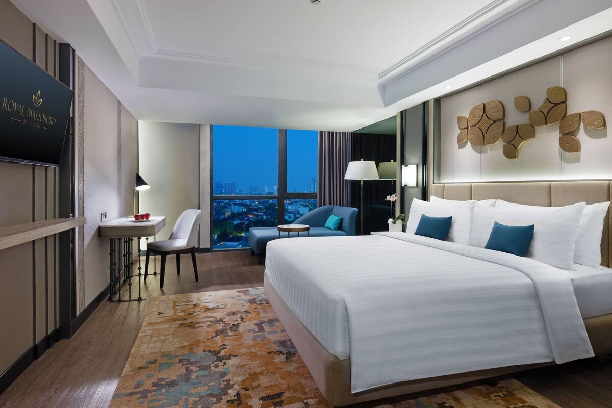 Ein Hotelzimmer mit einem großen Bett, einem Schreibtisch, einem Stuhl und Blick auf die Stadt im Abenddämmerung.