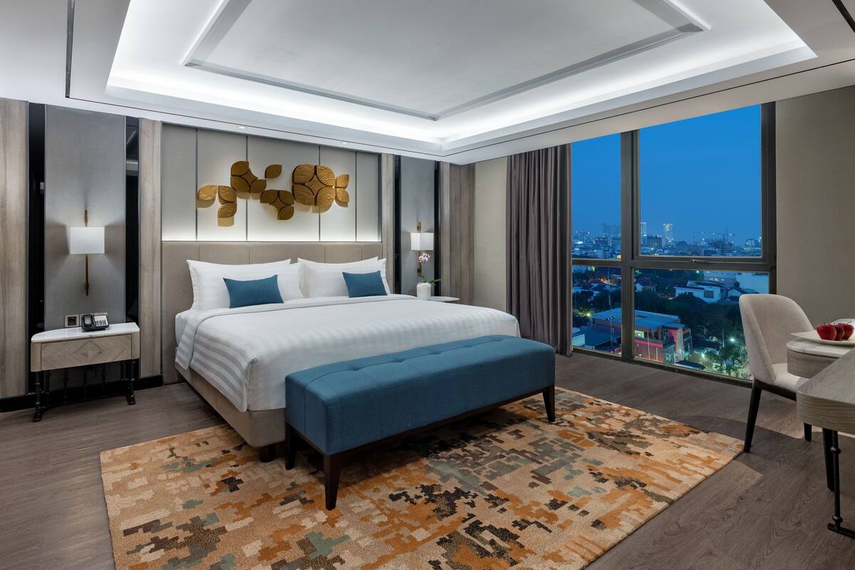 Ein modernes Hotelzimmer mit einem großen Bett, einem Schreibtisch und Blick auf die beleuchtete Stadt bei Nacht.