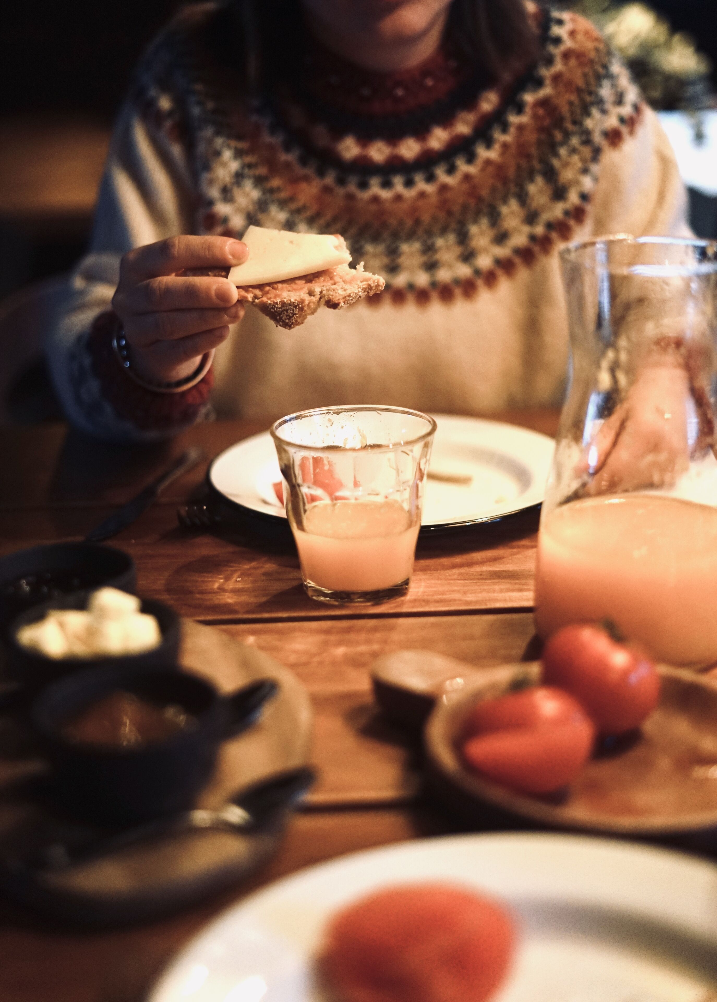 Una persona sosteniendo un trozo de pan con una capa de algo, un vaso de una bebida rosada y un vaso de un líquido más oscuro, posiblemente jugo, sobre una mesa de madera con platos y un tazón.