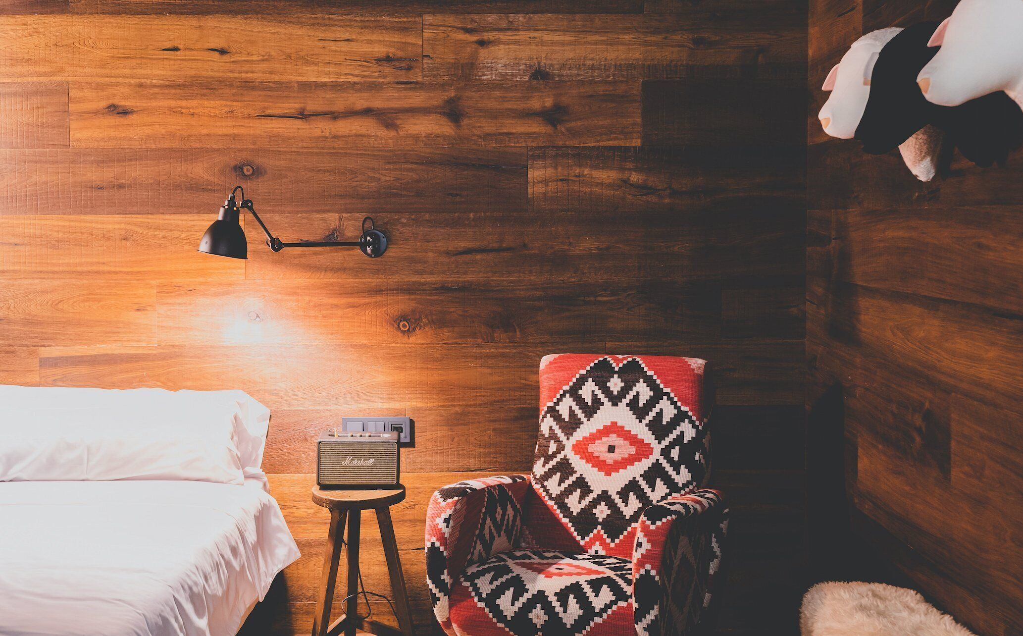 A cozy room with a wooden floor, a bed with white linens, a patterned armchair, a small stool with a speaker, and a lamp on the wall.