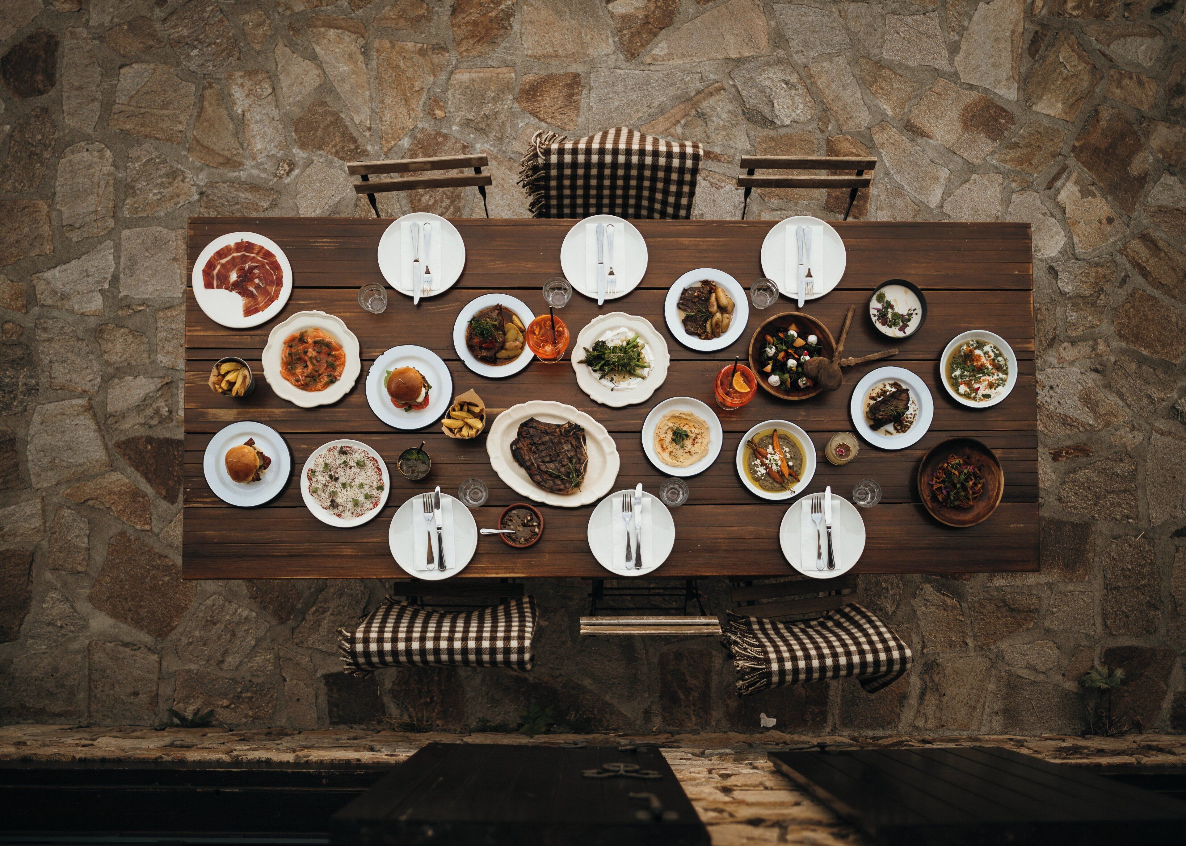 Una mesa de madera para comer dispuesta con diferentes platos y cubiertos frente a una pared de piedra.