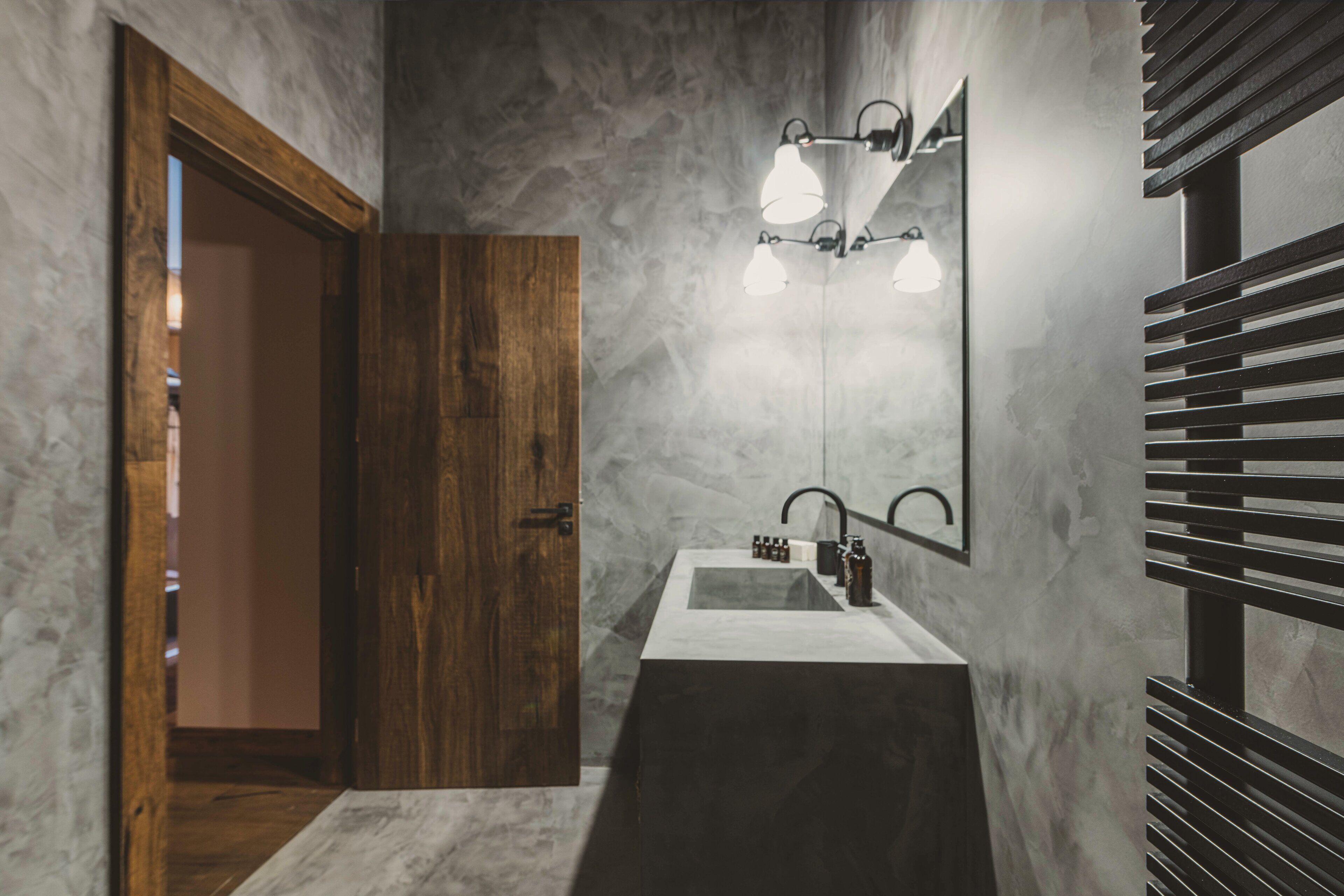 Un baño con puerta de madera, lavamanos blanco y espejo con luces.