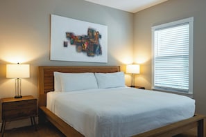 A bedroom with a wooden bed frame, white bedding, two bedside tables with lamps, a large abstract painting on the wall, and a window with blinds.