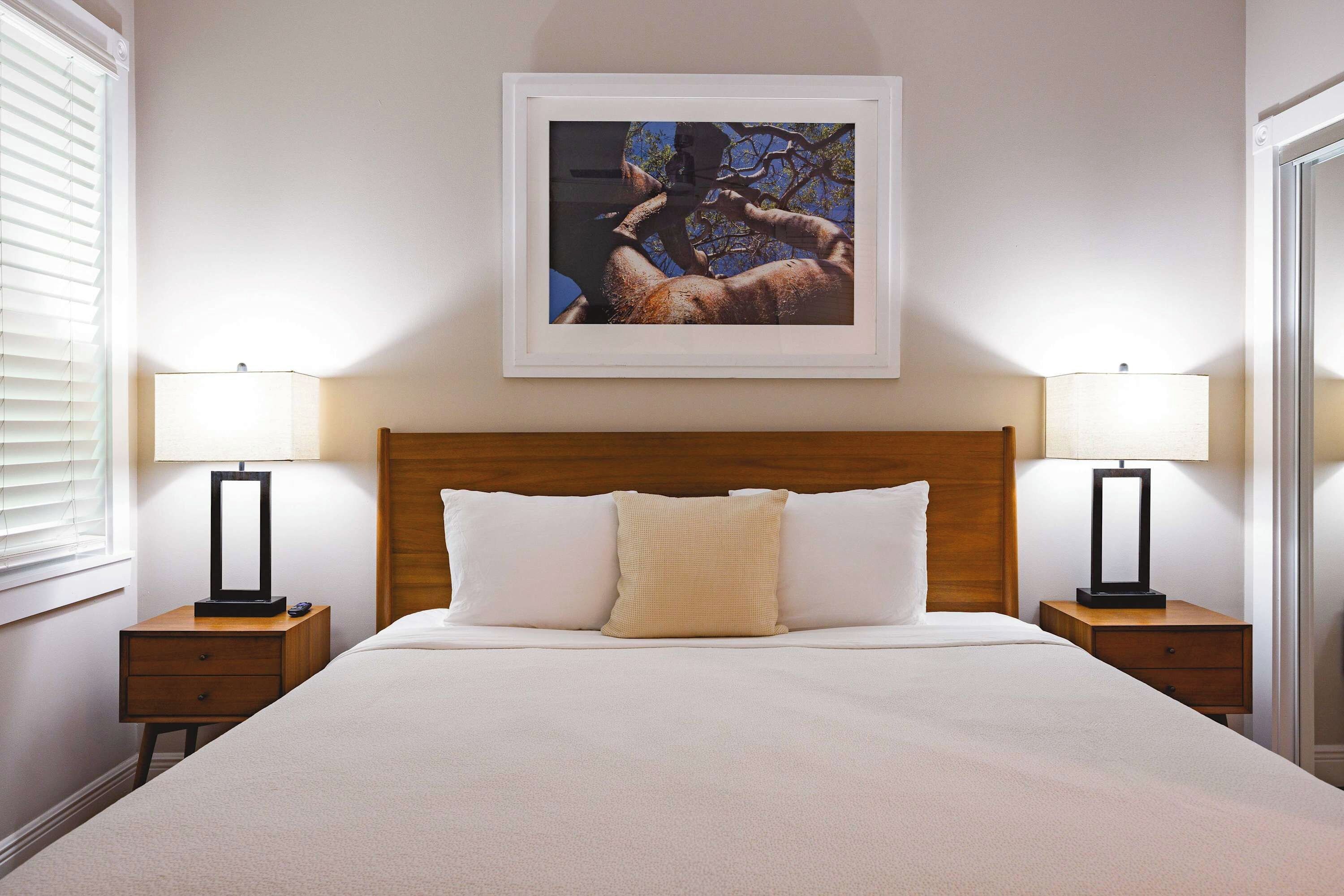 Une chambre d’hôtel avec un grand lit, deux tables de chevet avec des lampes, un tableau encadré au mur et une fenêtre avec des stores.