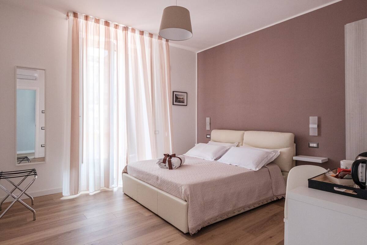Une chambre moderne avec un grand lit, une table de chevet, une lampe et une vue sur l’extérieur à travers des rideaux légers.