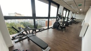 A gym with various exercise equipment, large windows, and a view of the city. - Moov'Appart Hotel Clichy (Clichy)