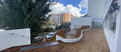 A rooftop terrace with wooden flooring, a curved railing, and a view of a parking area and buildings in the background.