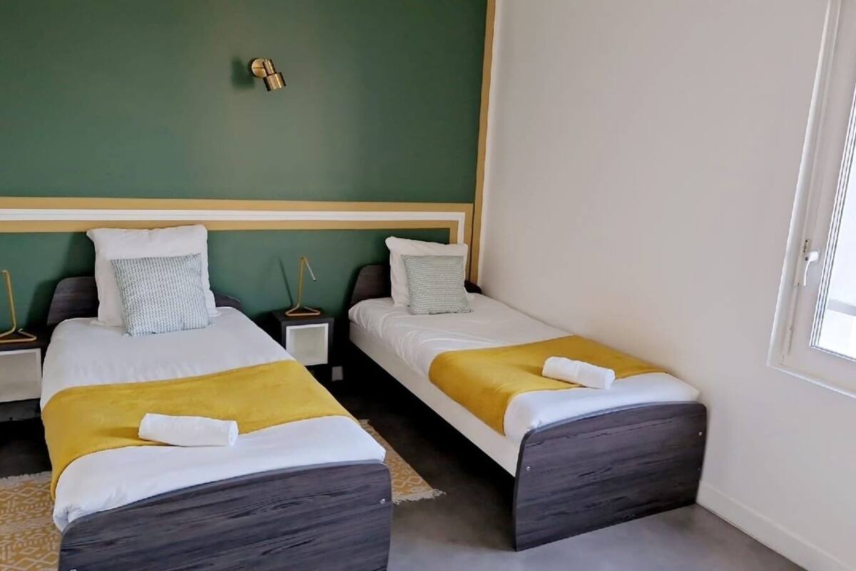 A hotel room with two single beds, each with a yellow blanket and white pillows. There is a bedside table with a lamp on each bed. The room has a green accent wall and a window on the right side.