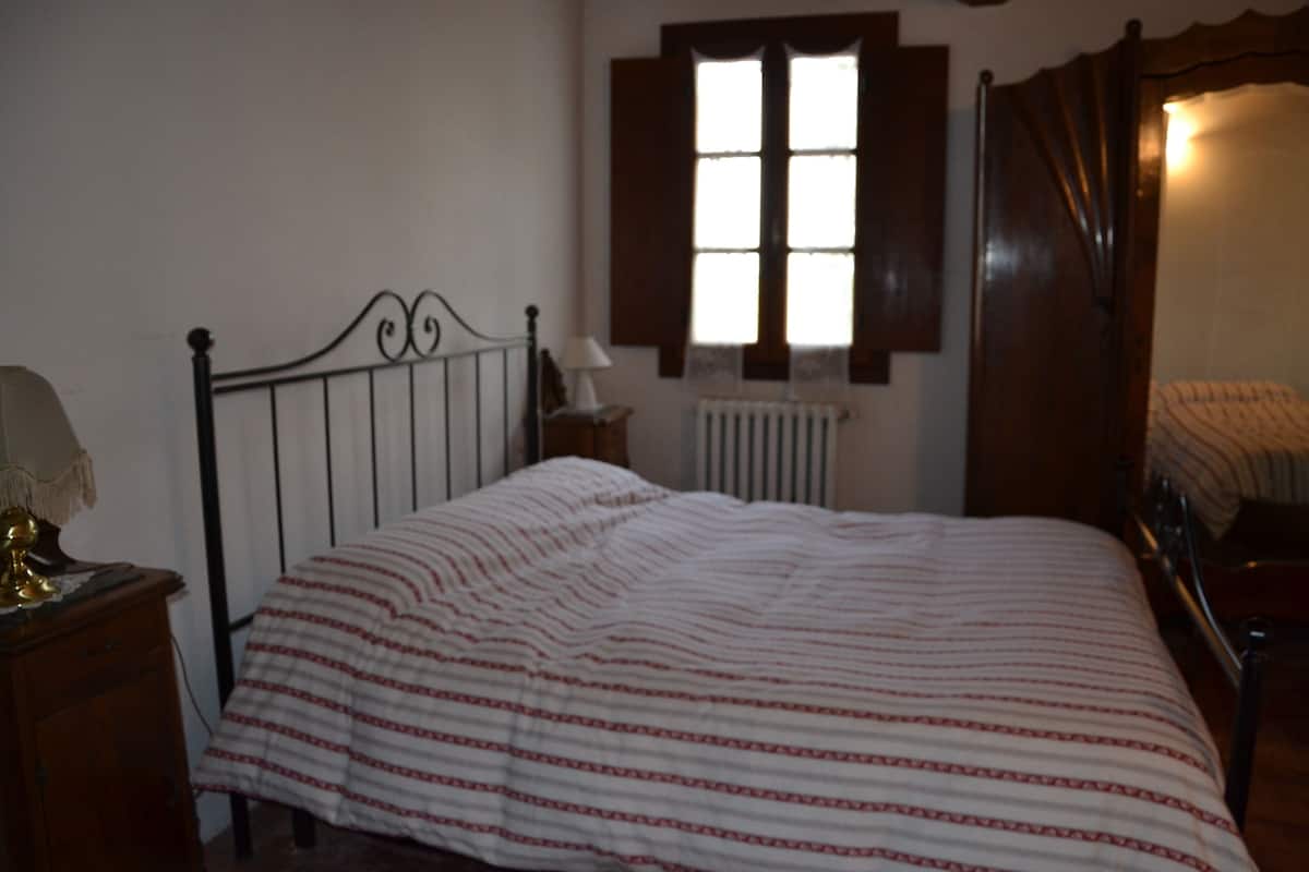 A bedroom with a bed, a nightstand, a lamp, a window with shutters, and a radiator.
