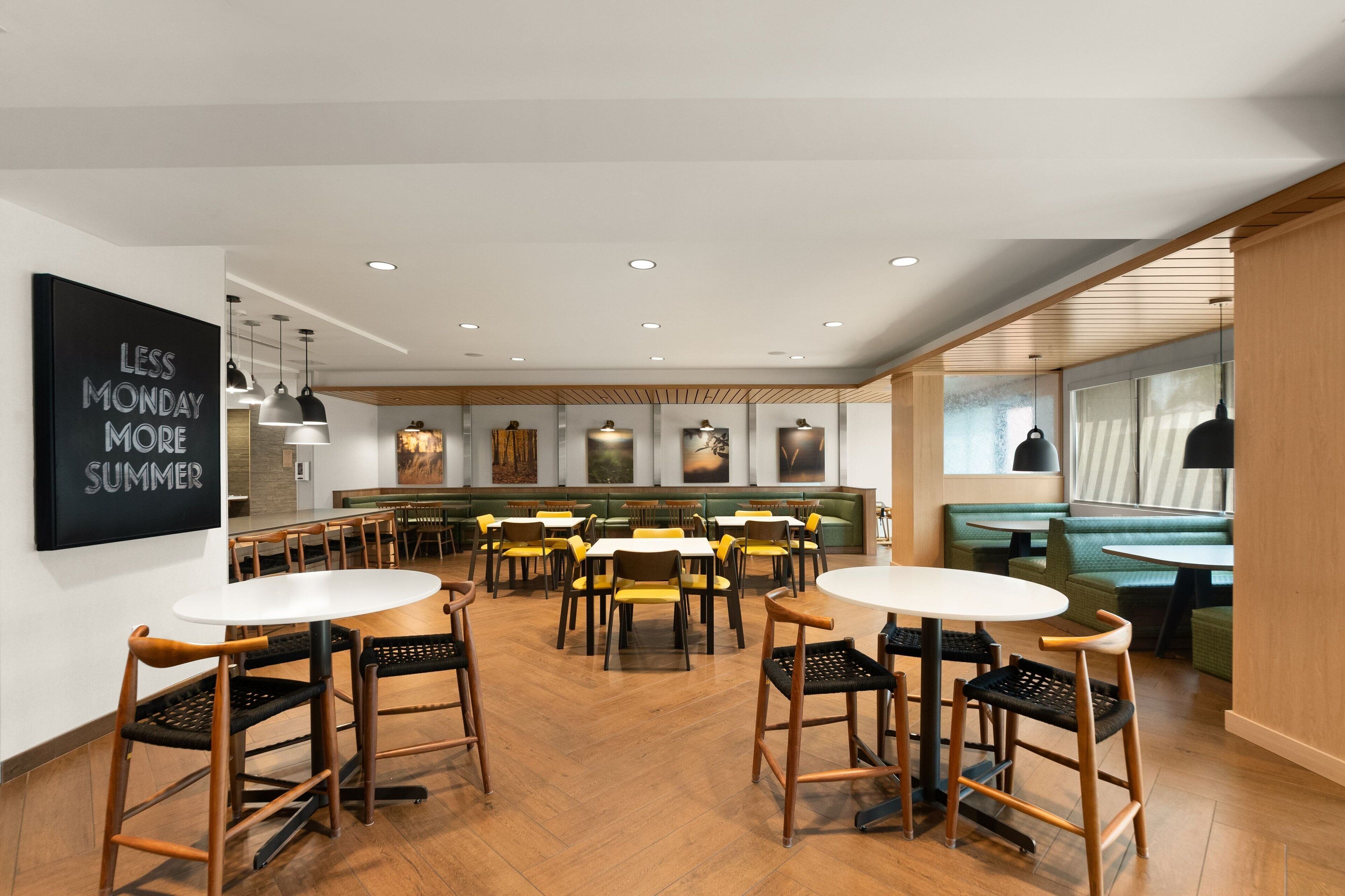 Un'area ristorazione moderna con tavoli e sedie in legno, un grande cartello nero sulla parete e vista sull'area bar sullo sfondo.