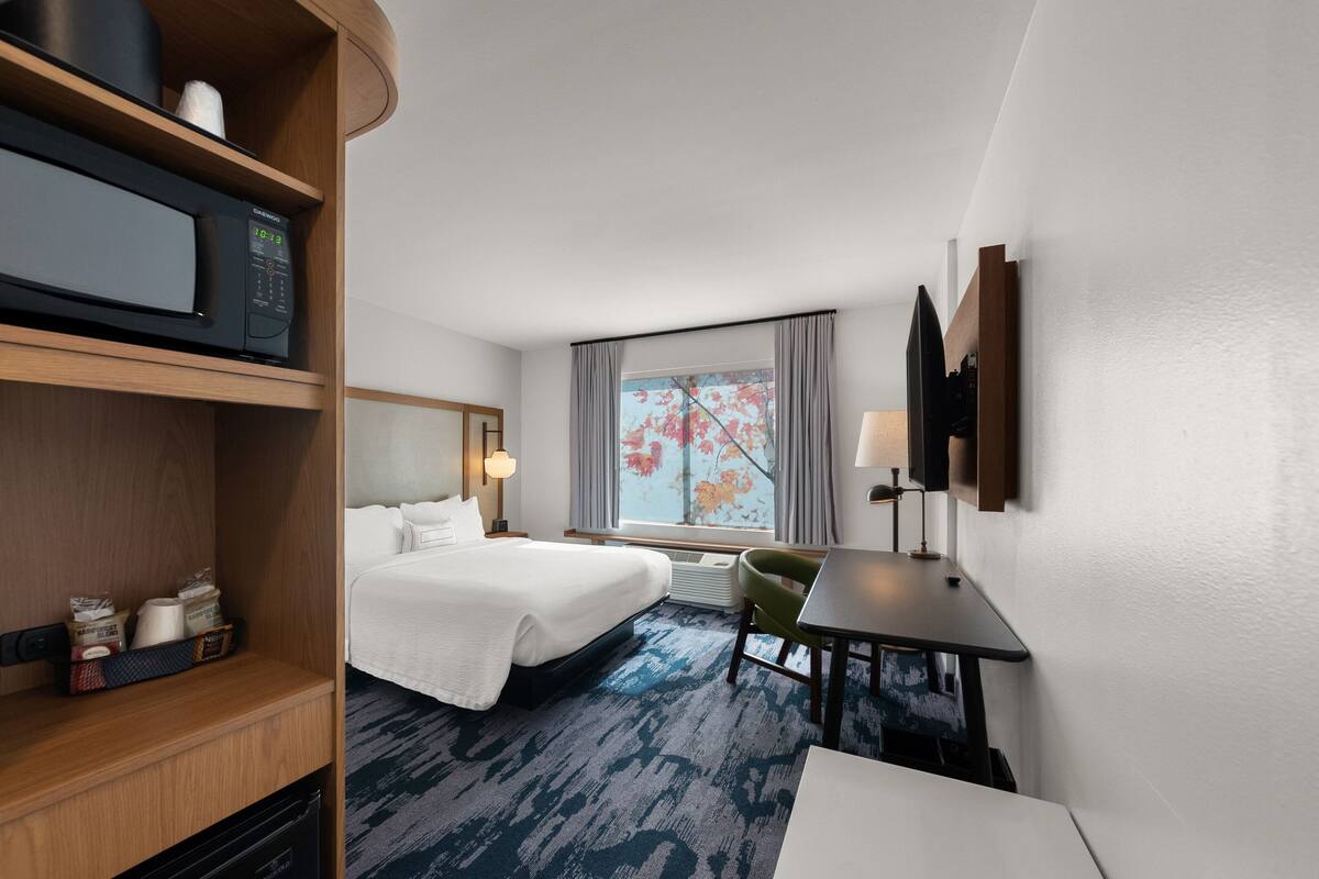 A hotel room with two beds, a desk, a television, a window with curtains, and a microwave on a shelf.
