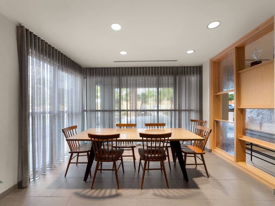 Uma área de refeições com mesa e cadeiras de madeira, janelas amplas com cortinas e uma estante de livros de madeira.