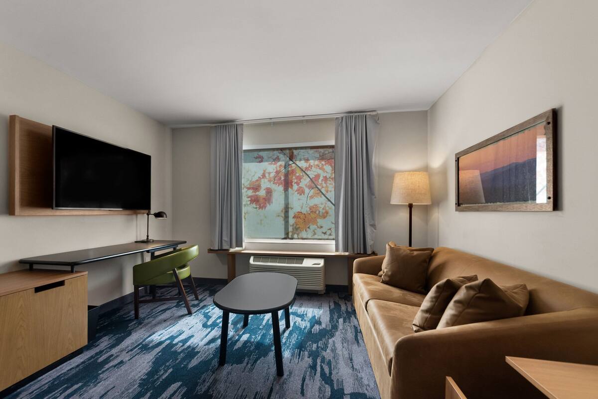 A hotel room with a flat-screen TV, a desk, a green chair, a brown sofa, and a window with curtains.
