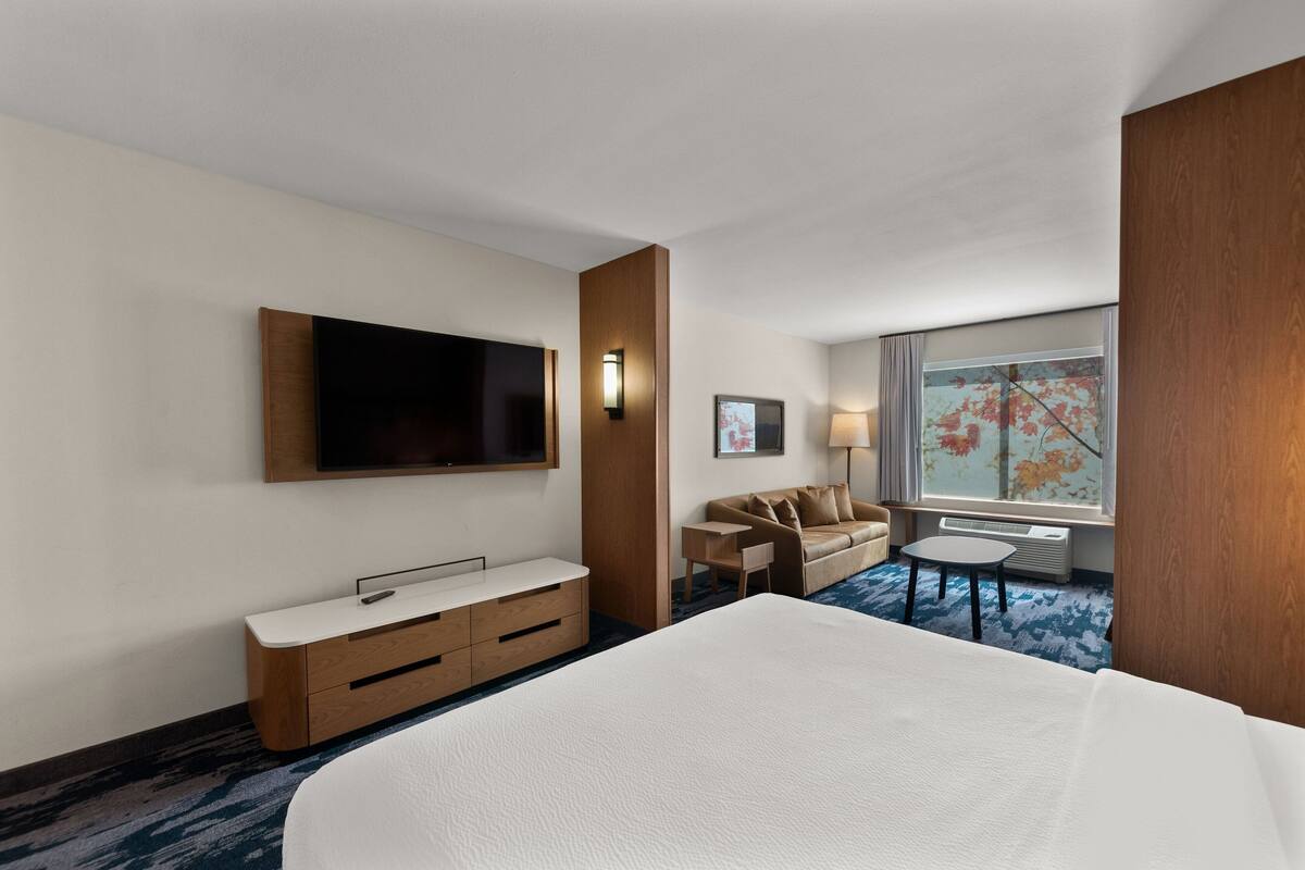 A hotel room with a large bed, a TV mounted on the wall, a sofa, a small table, and a window with a view of trees.