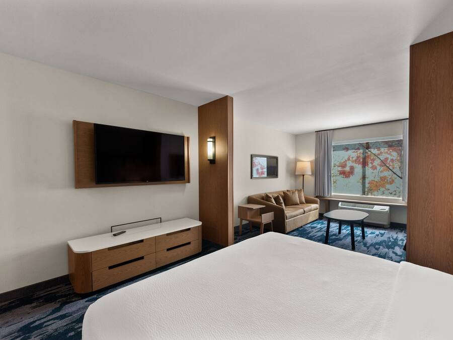 A hotel room with a large bed, a TV mounted on the wall, a sofa, a small table, and a window with a view of trees.
