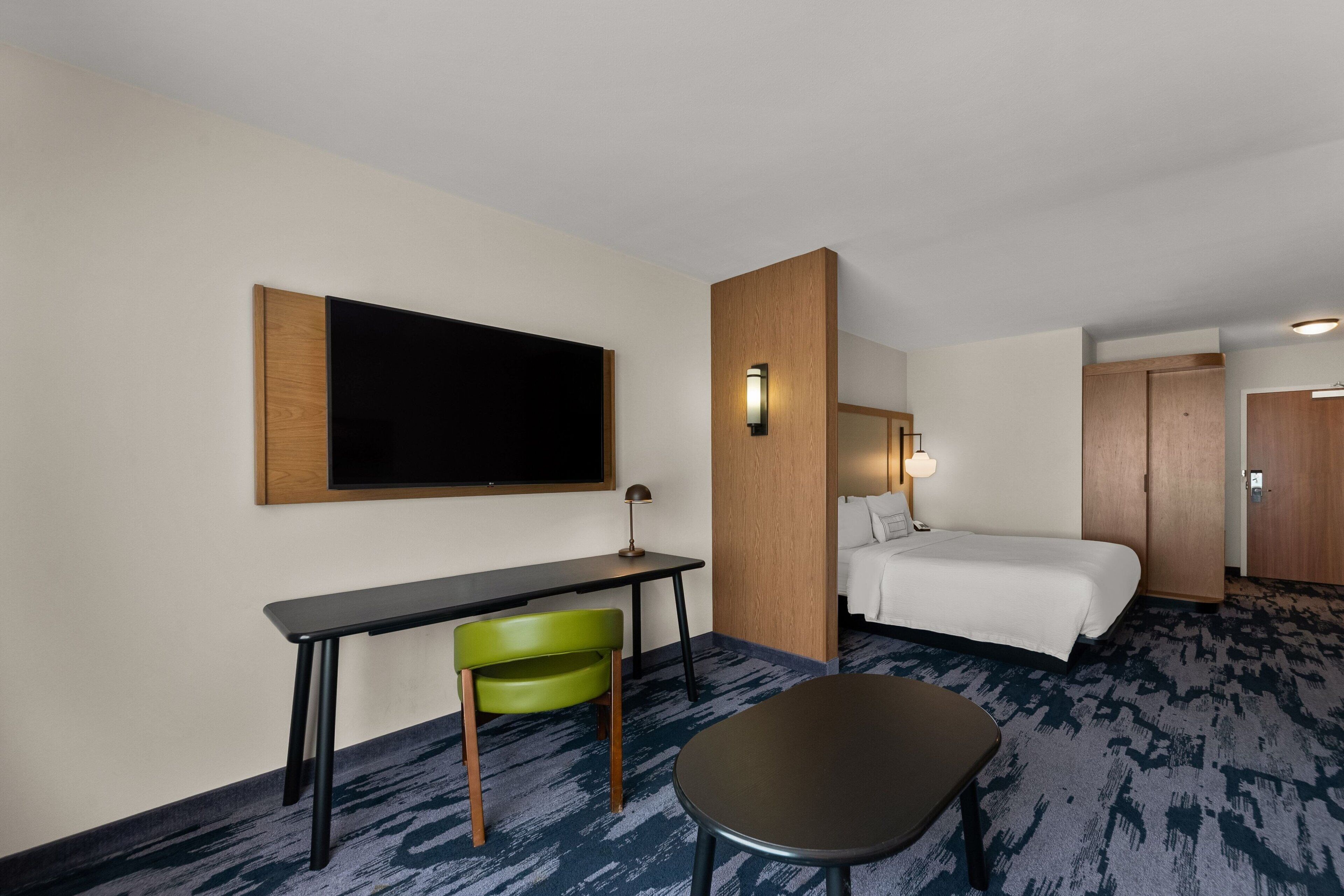 A hotel room with a bed, a desk with a chair, a television, and a carpeted floor.