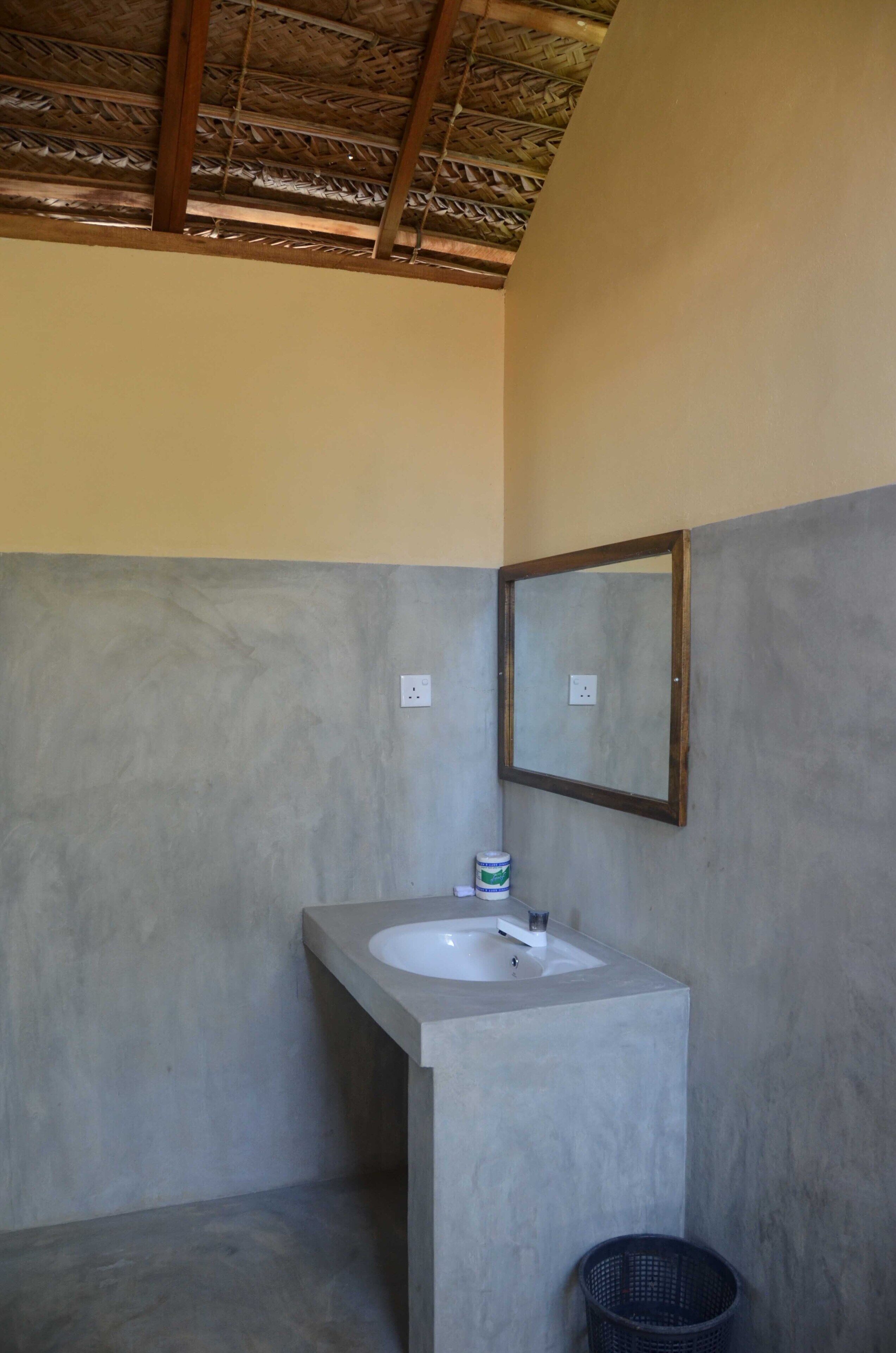 Un baño con un lavamanos de concreto, un espejo con marco de madera y un techo de paja.