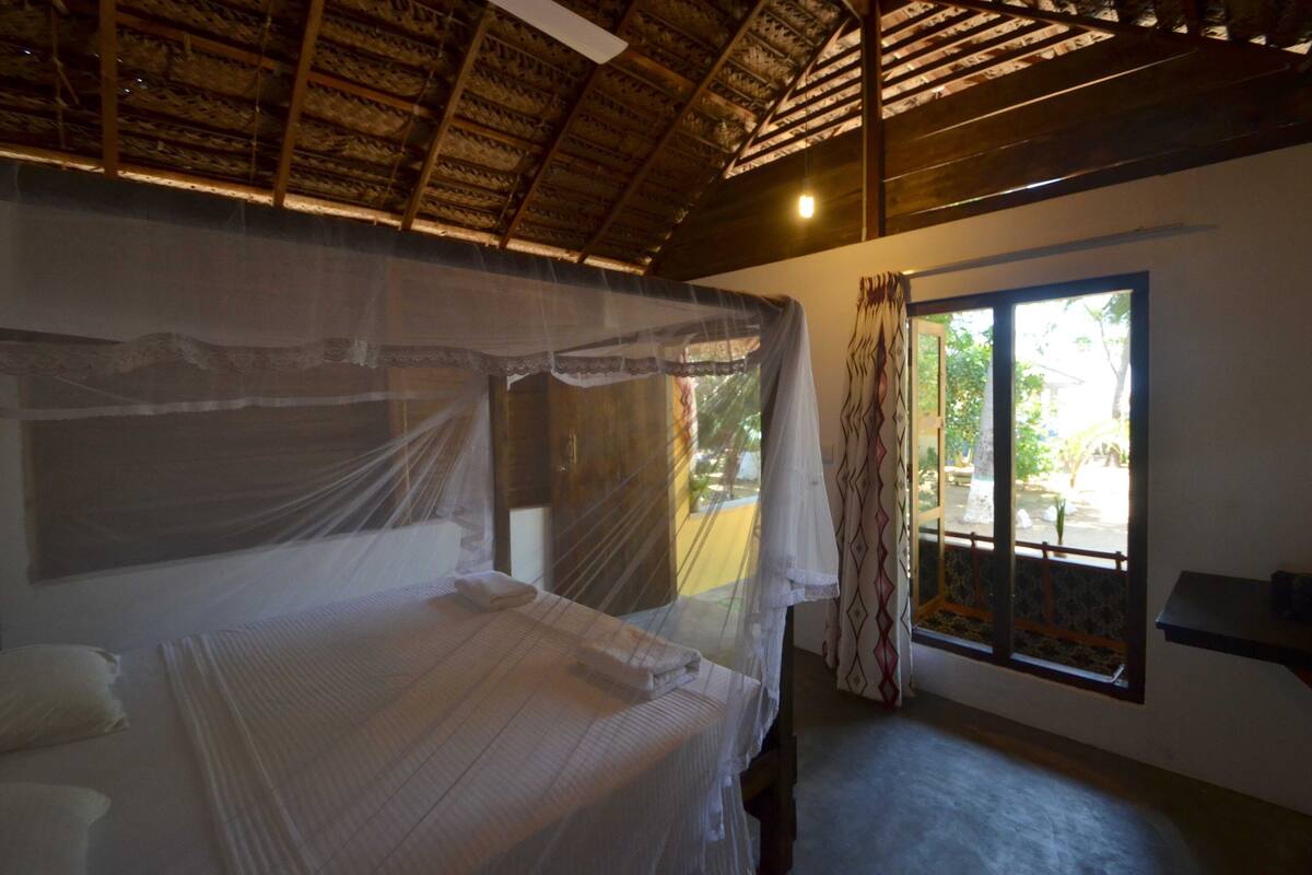 A room with a bed covered by a mosquito net, a window with a view of the outdoors, and a ceiling with wooden beams.