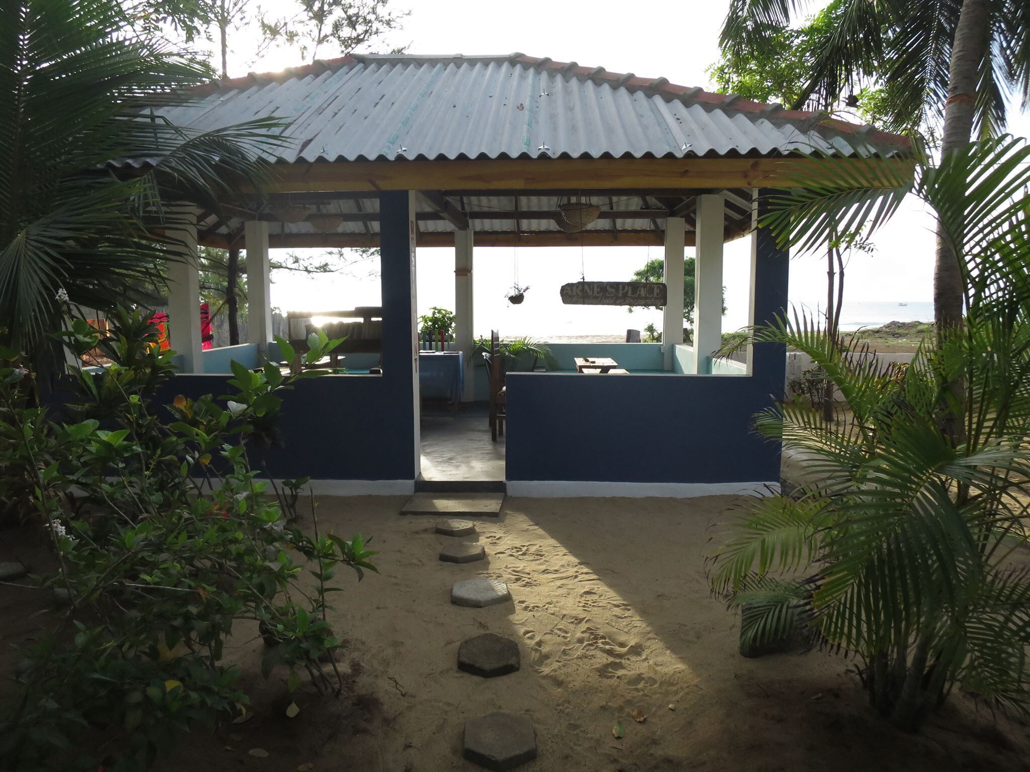 A covered outdoor seating area with a view of the beach, featuring a wooden table and chairs, surrounded by plants and a sandy path.